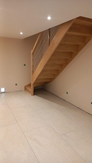 Intérieur d'une pièce avec un escalier en bois et murs clairs, carrelage au sol.